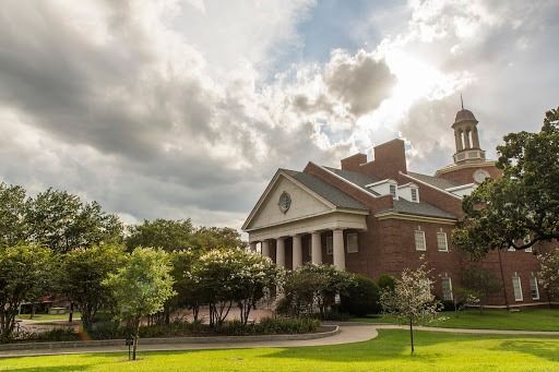 Brick building with columns, cupola, and trees on a green lawn under a cloudy sky.