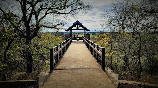 Wooden bridge with a covered viewpoint over a landscape of trees under a cloudy sky.