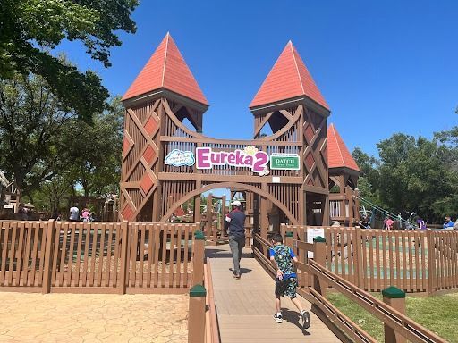 Wooden playground with towers and archway; Eureka 2 sign. People walking. Sunny day.