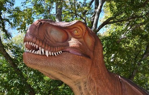Brown T-Rex head with closed eyes, outdoors in front of green trees.