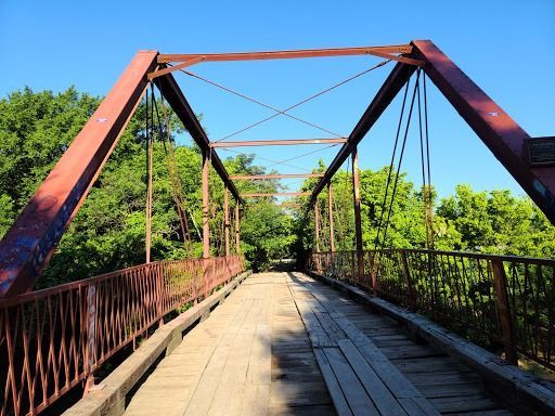 Red metal truss bridge over a wooded area with a wooden walkway, under a blue sky.