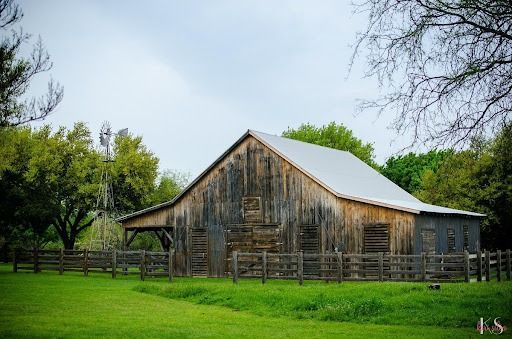 Weathered wooden barn with a metal roof, surrounded by a wooden fence and green grass under a cloudy sky.