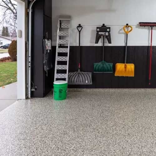 Garage interior with gray speckled floor, tools hanging on a wall, ladder, and open door to yard.