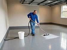 Man applying epoxy coating to a gray garage floor. He is using a sprayer near a bucket.