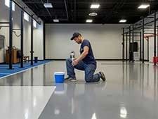 Man on one knee, cleaning gray glossy floor in an empty gym. White walls and equipment.