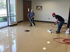 Two men cleaning a shiny beige floor in a room with windows and a door; they use vacuums.