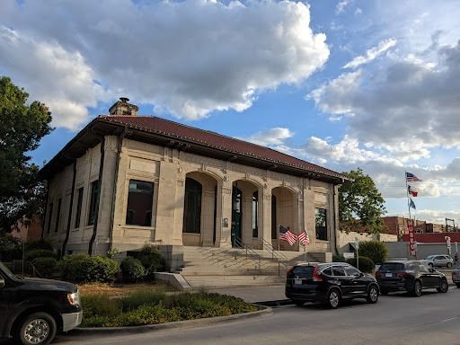 Beige stone building with arched windows, red tile roof, and American flags on a sunny day.