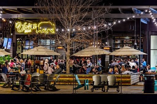 Night scene, outdoor restaurant patio with people dining. Lit by string lights and a neon sign.