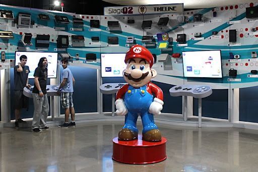 Mario statue stands in a gaming exhibit, with people looking at displays of past consoles.