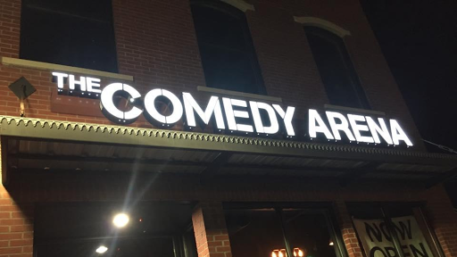 Exterior of The Comedy Arena at night with a lit-up sign against a brick building.
