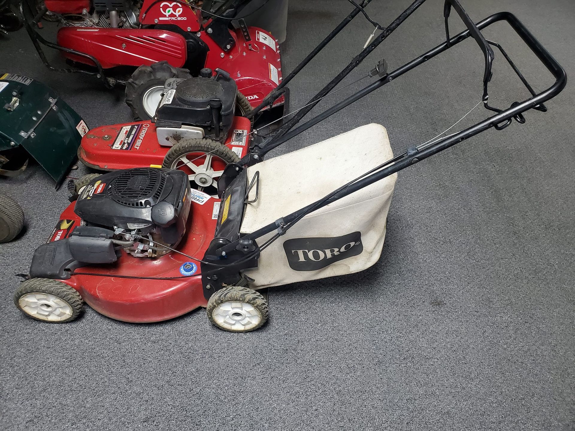 A red lawn mower with a white bag is sitting on a carpeted floor.
