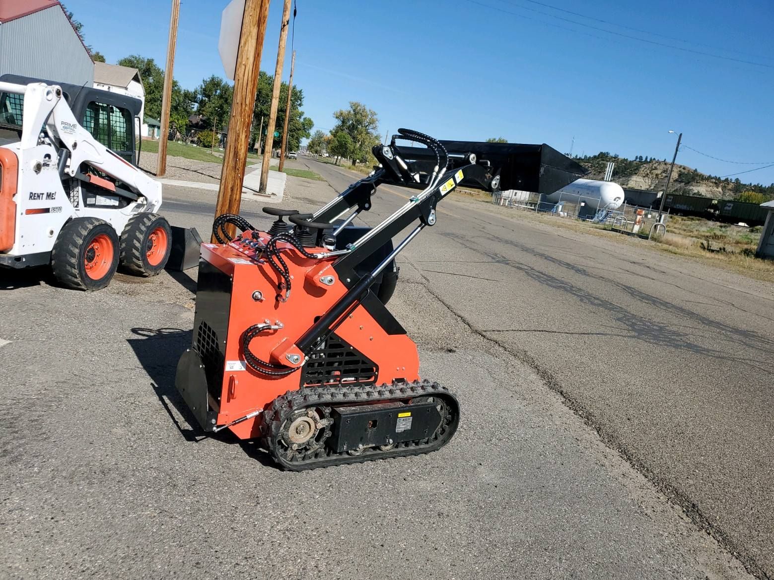 A small tractor is parked on the side of the road next to a bobcat.