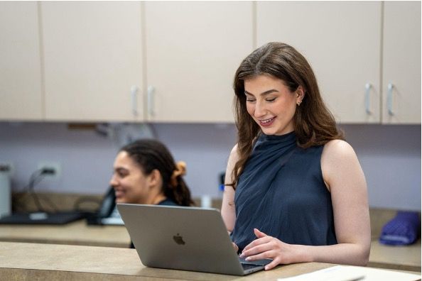 Person using a laptop at a table, with another person in the background, in a classroom or office setting.