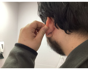 A person’s hand pulls on their earlobe while looking toward a mirror in a room with a wall-mounted soap dispenser.