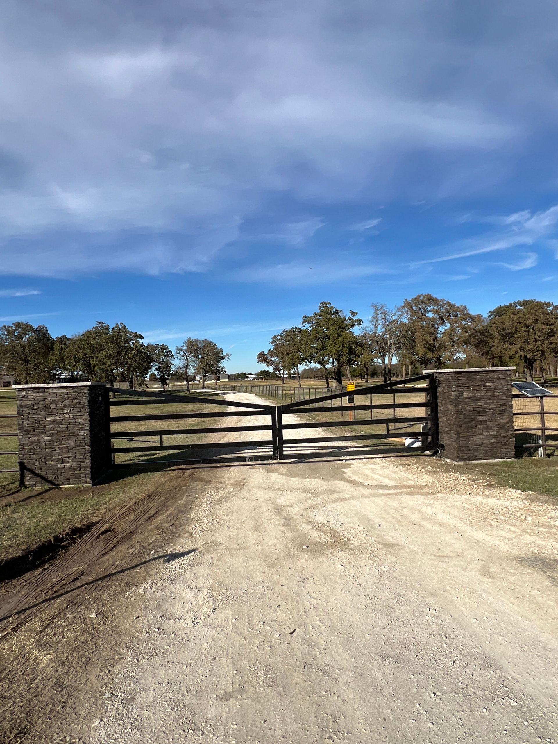 Gated gravel driveway leading to a ranch with brown wooden gates and stone pillars, under a cloudy sky.