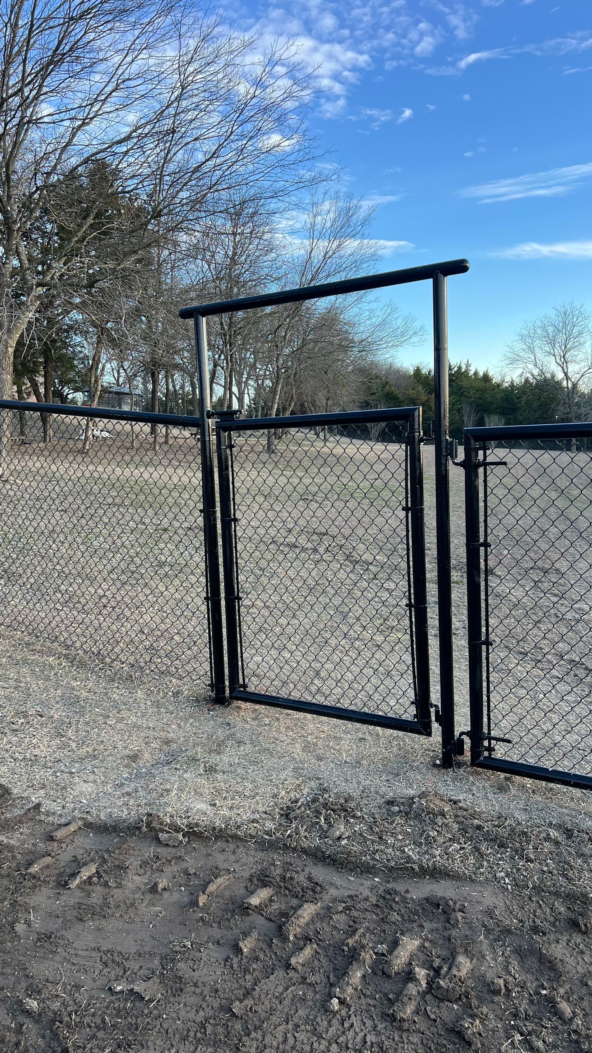 Black chain-link fence and gate in gravel. Overcast sky and bare trees in the background.