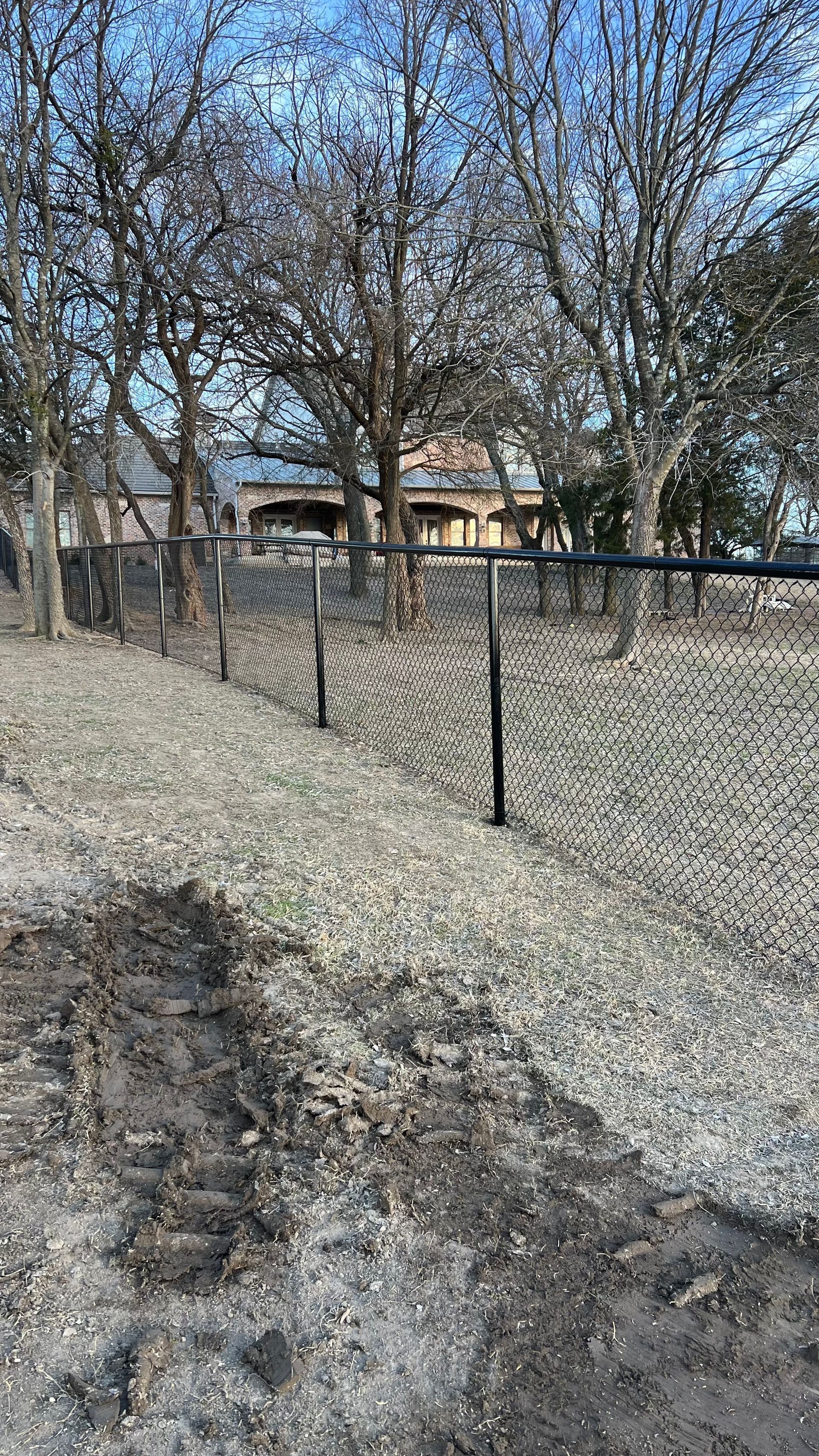 Black fence with black posts in a gravel area, trees in the background, daytime.