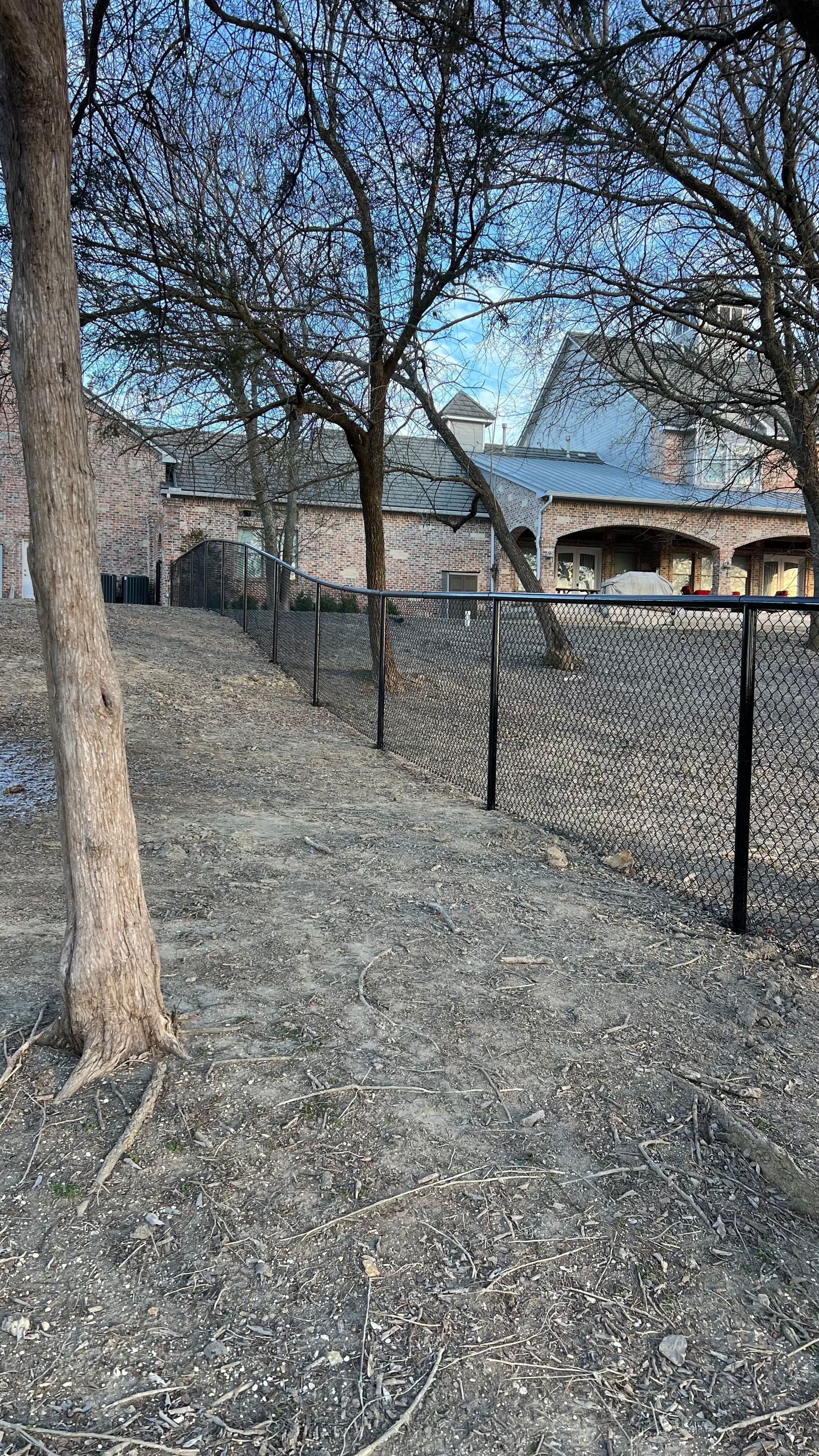 Chain-link fence on a gravel pathway between trees, leading toward a building with a blue sky background.