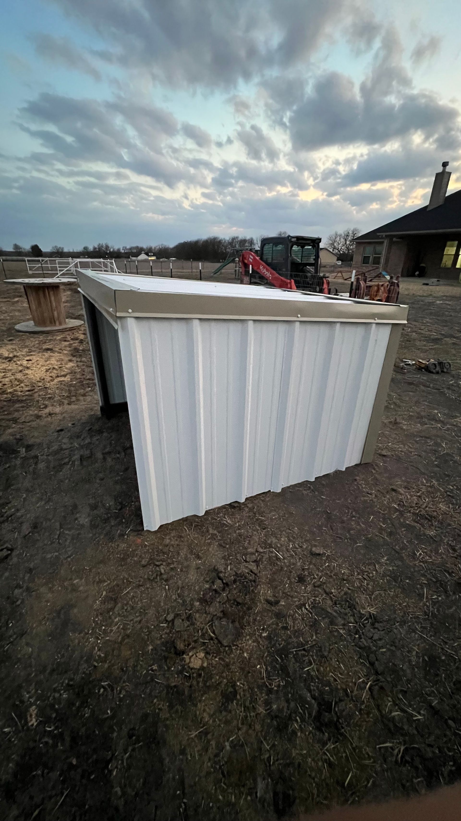 White metal shed under construction on a dirt lot, with equipment and a cloudy sky.