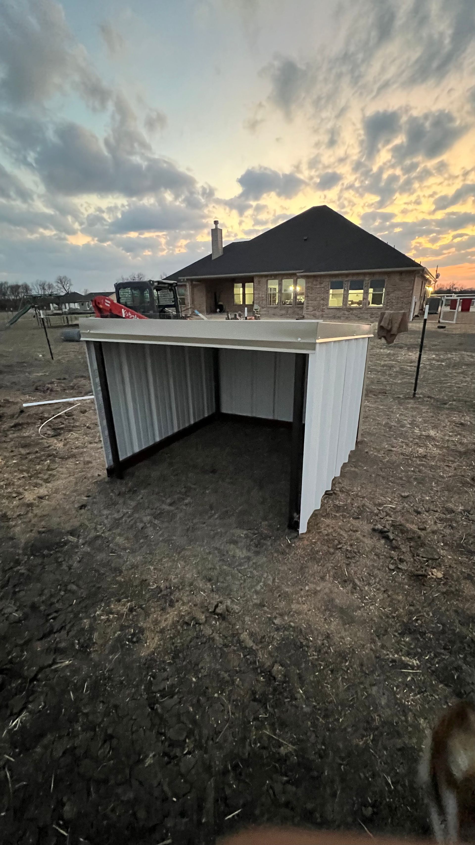 White and black wooden shed in a grassy field, house in the background at sunset.