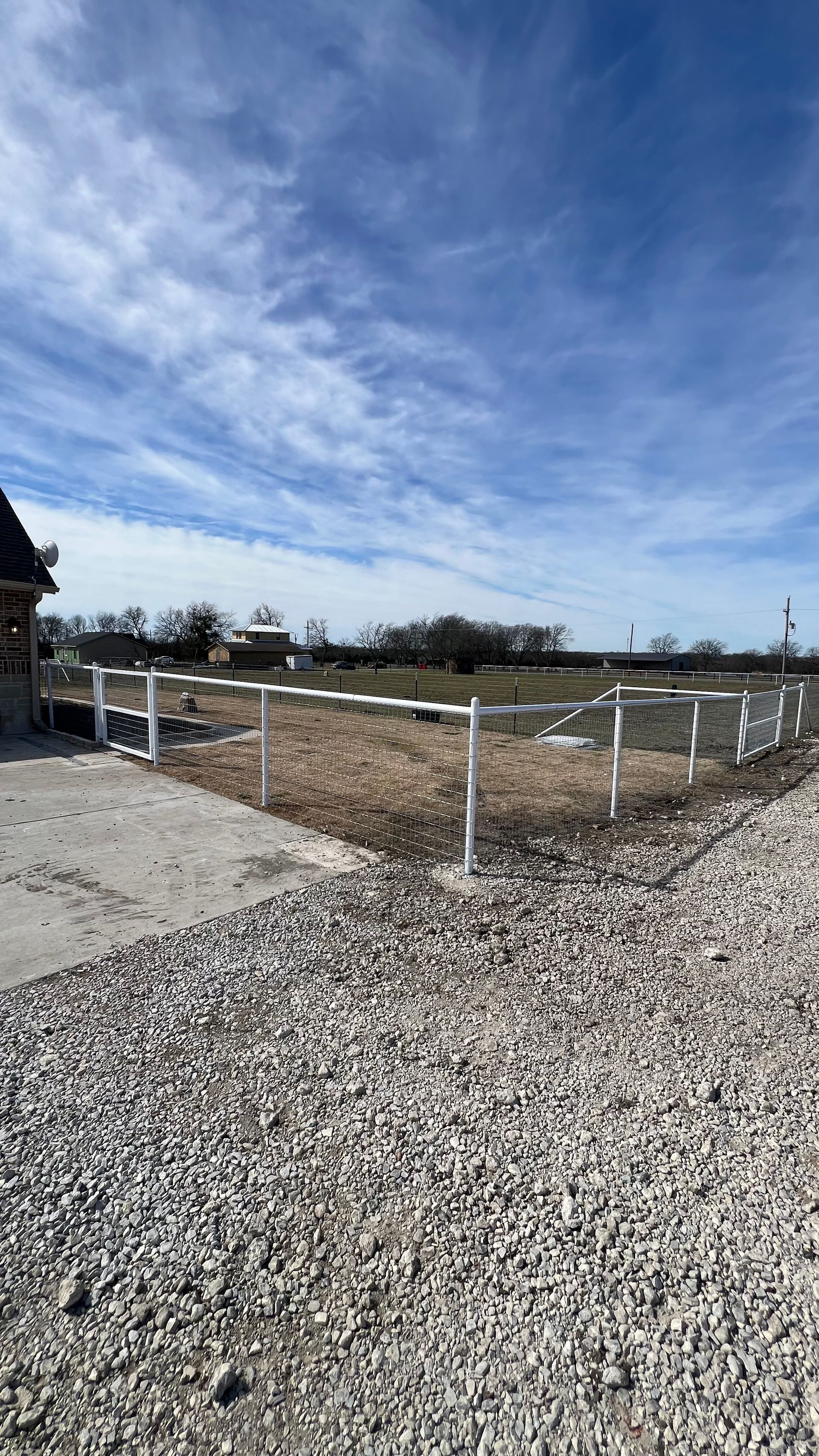 Gravel driveway leads to a white fenced-in area with a partly cloudy blue sky in the background.