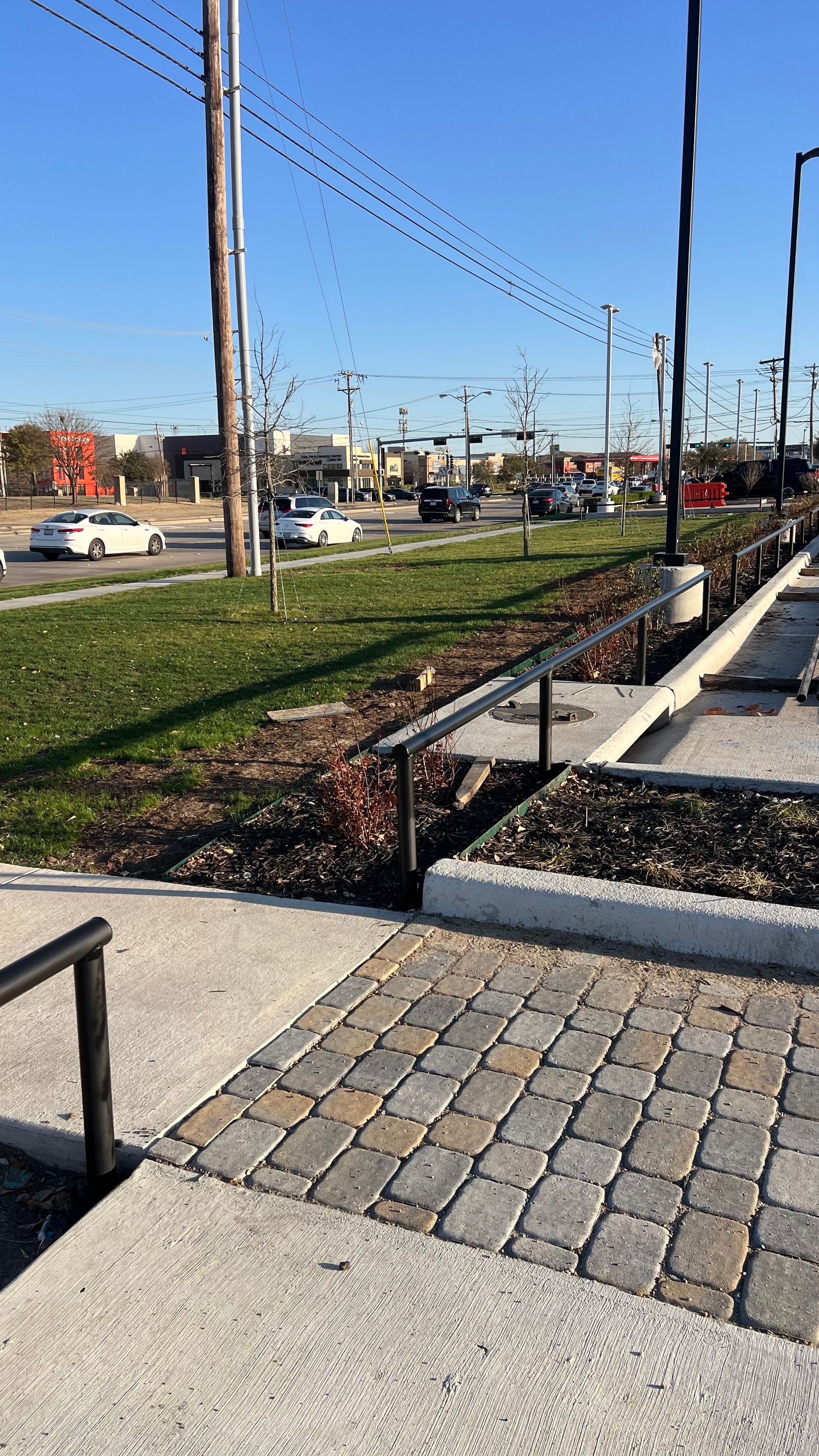 View of a grassy median, brick walkway, and street with traffic. Sunny day.