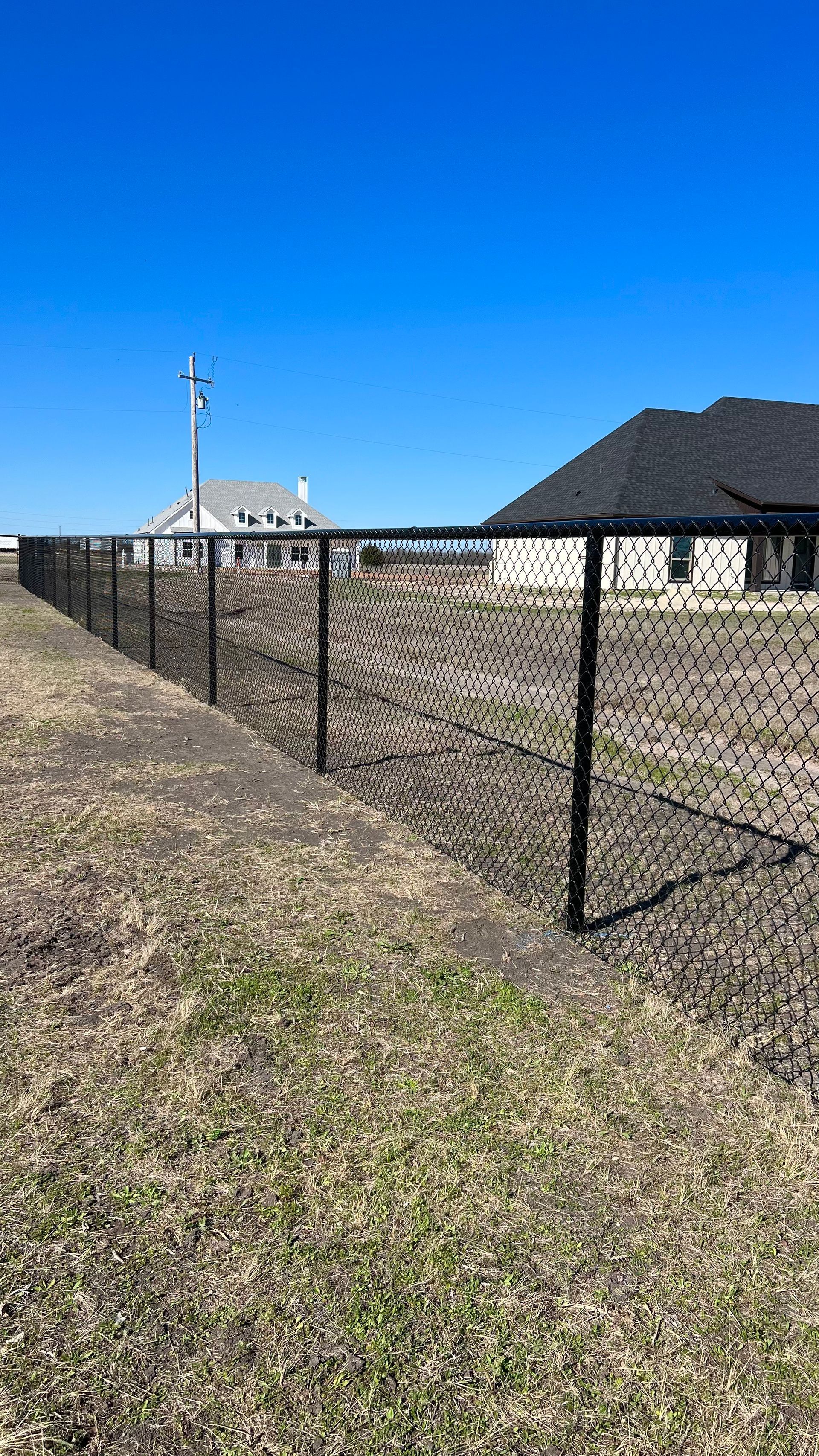 Black chain-link fence bordering a grassy field, under a clear blue sky. Houses in the background.