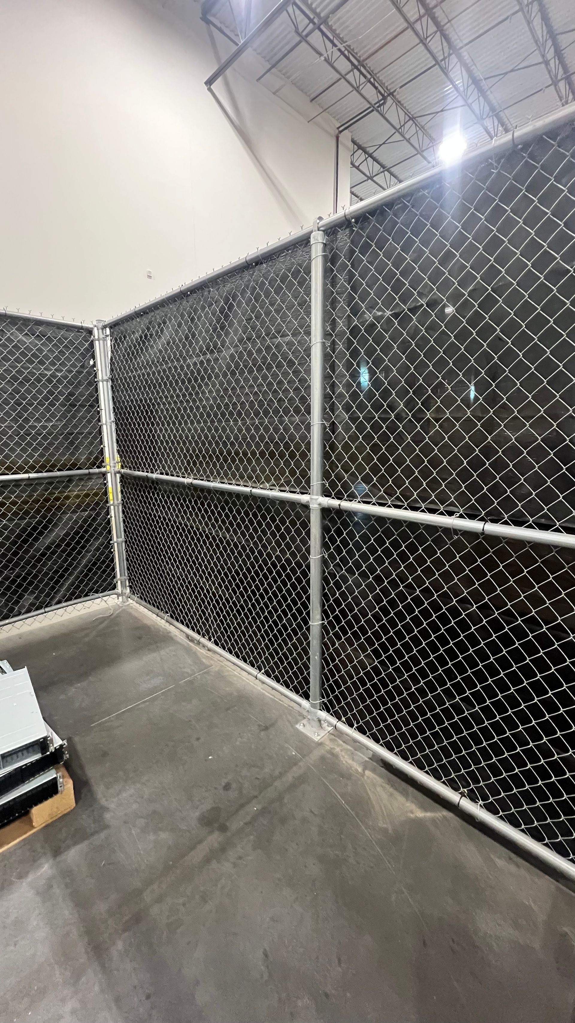 Chain-link fence with black privacy slats inside a warehouse. Concrete floor, metal framing.