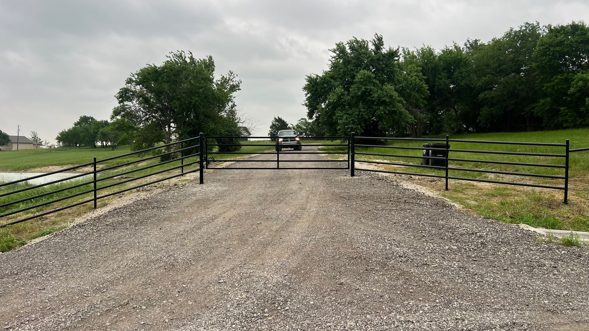 Gravel driveway leading to a closed black gate. A car is visible further down the drive with trees in the background. Overcast sky.