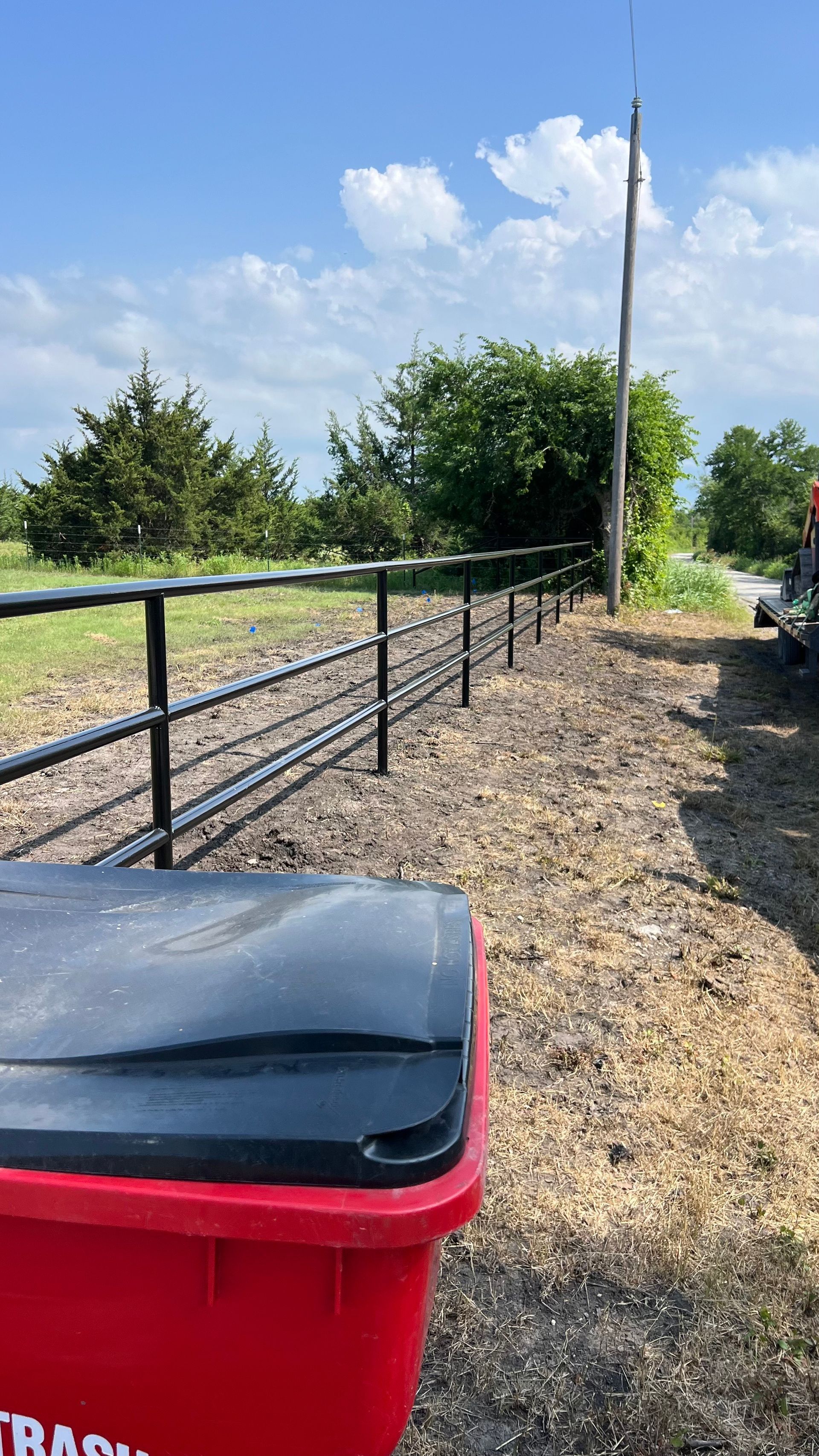Red trash bin in foreground, black metal fence extends into distance along a gravel road, sunny day.