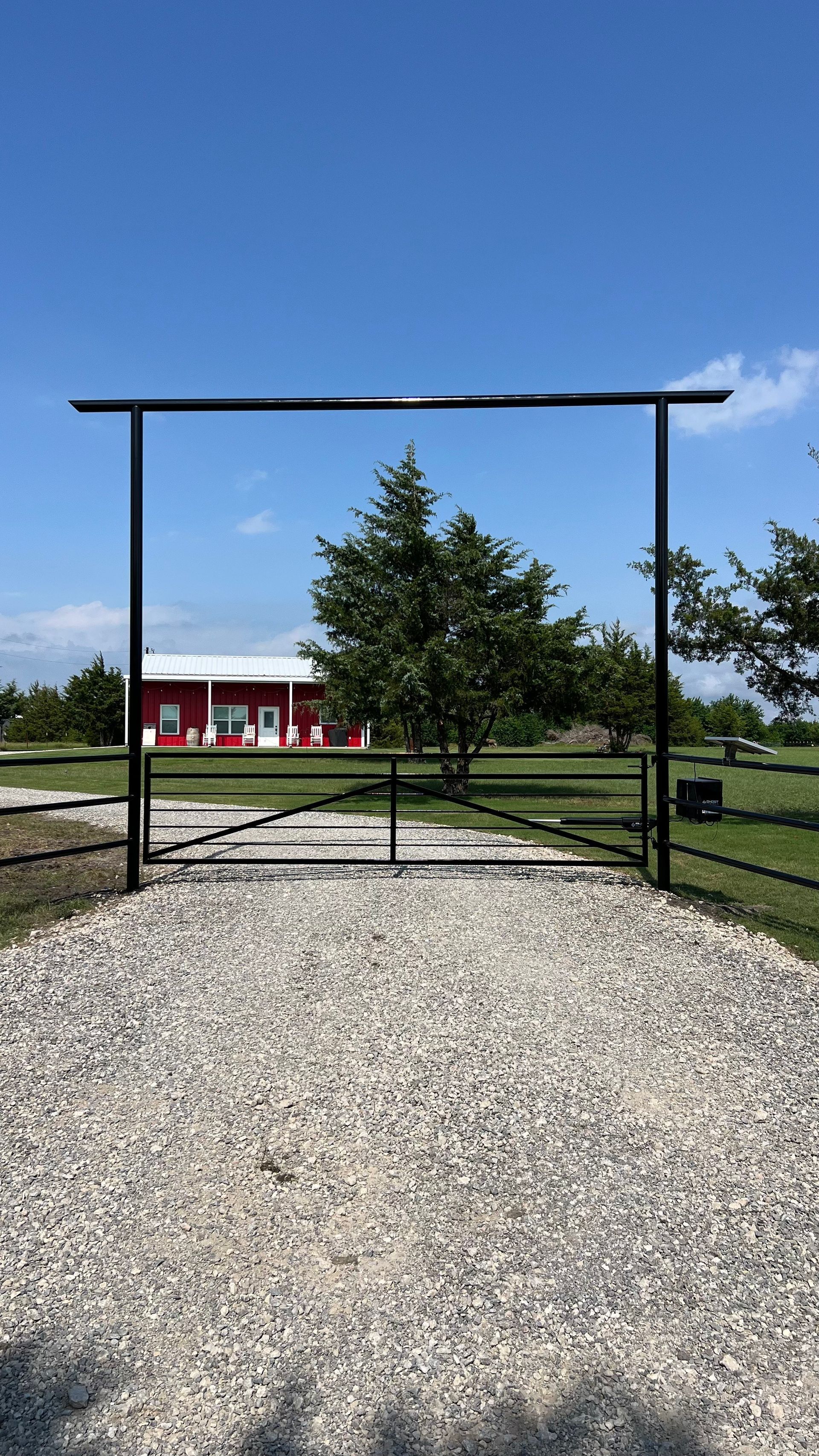 Black metal gate and archway on a gravel driveway, leading to a red building on a grassy property.