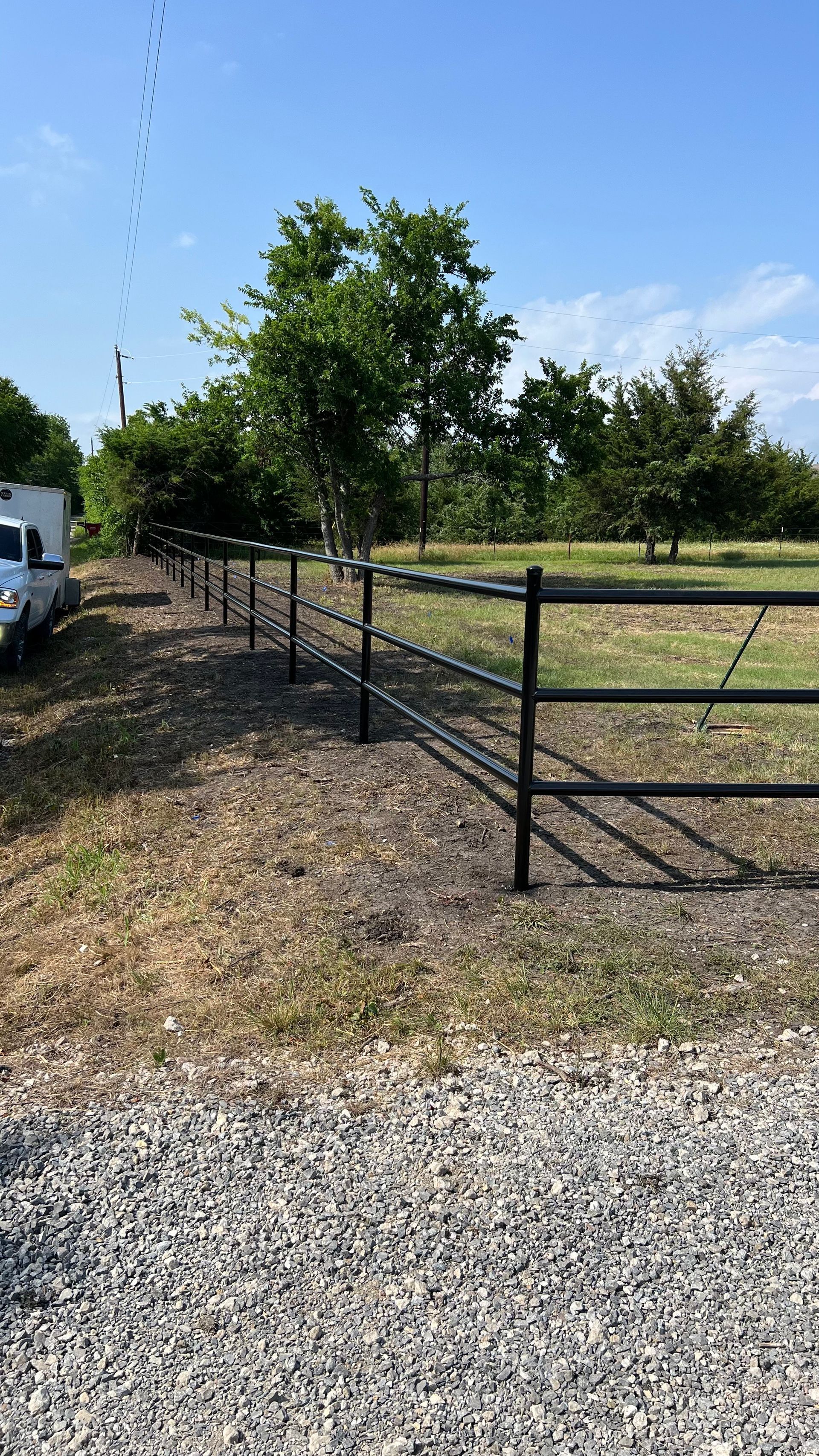 Black fence in front of a tree line on a sunny day with gravel ground and a bright blue sky.
