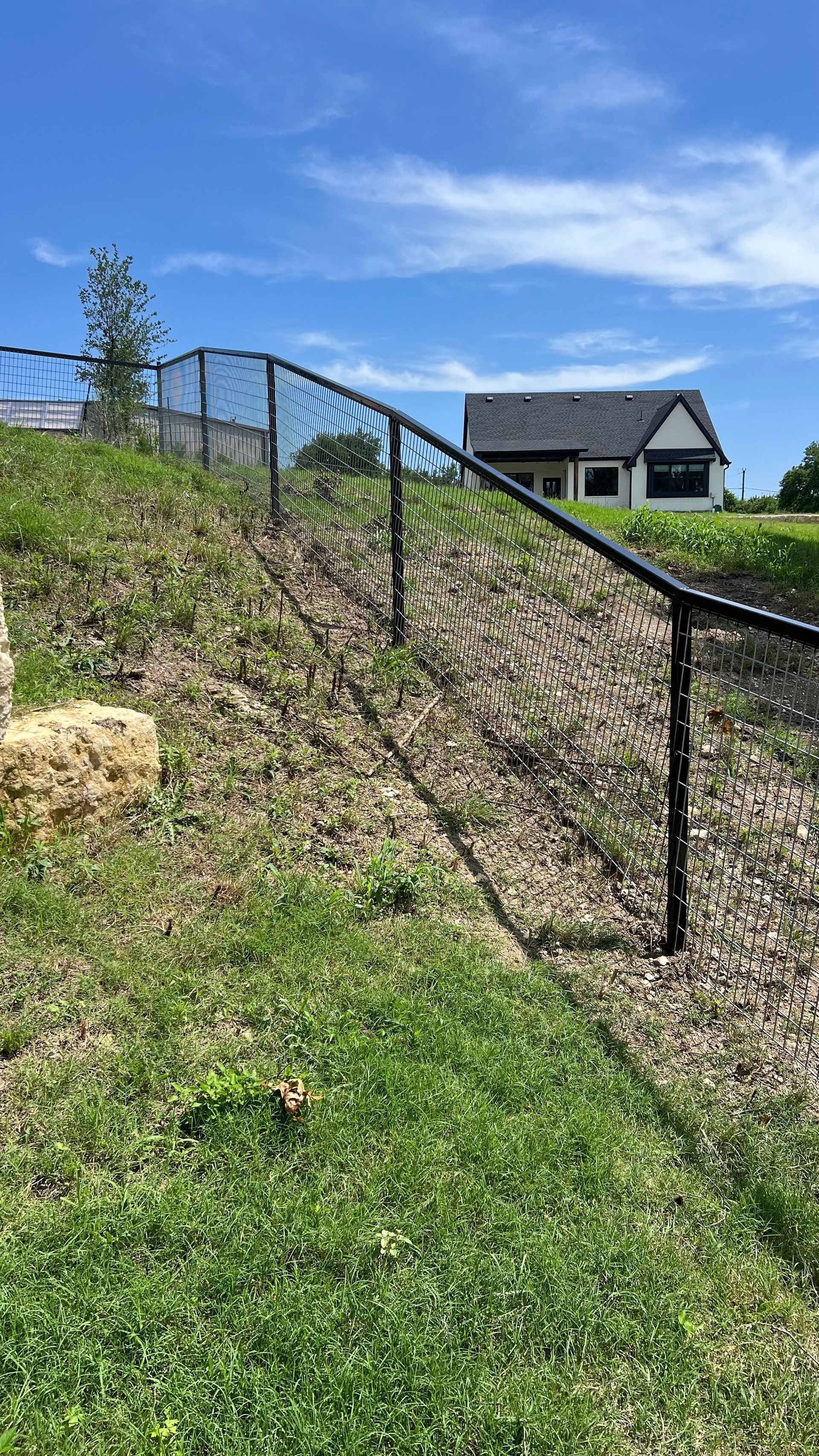 Black metal fence on a grassy hill, leading to a house under a blue sky.
