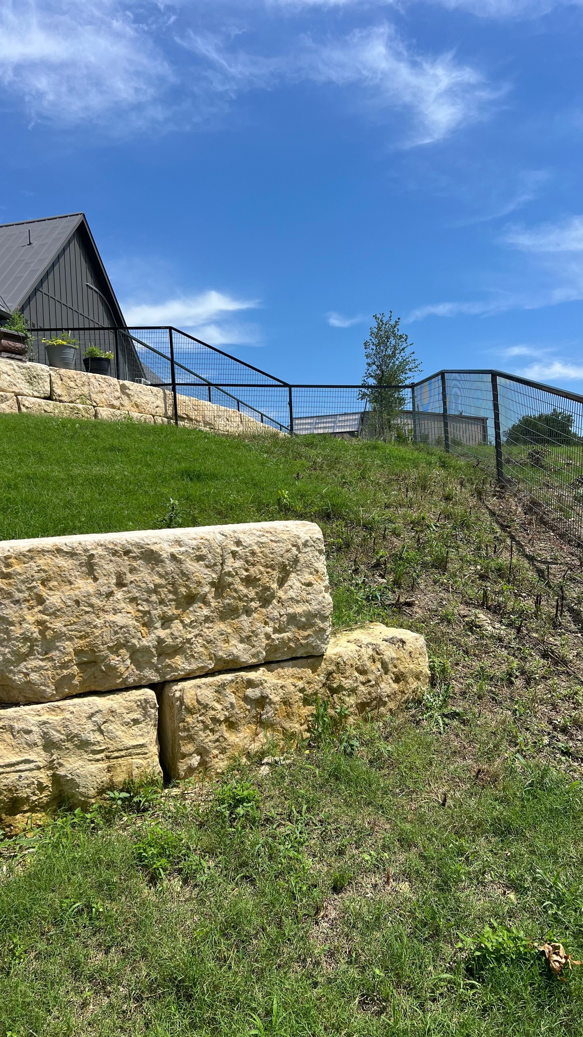 Stone retaining wall with grassy hill, black metal railing, and a house under a blue sky.