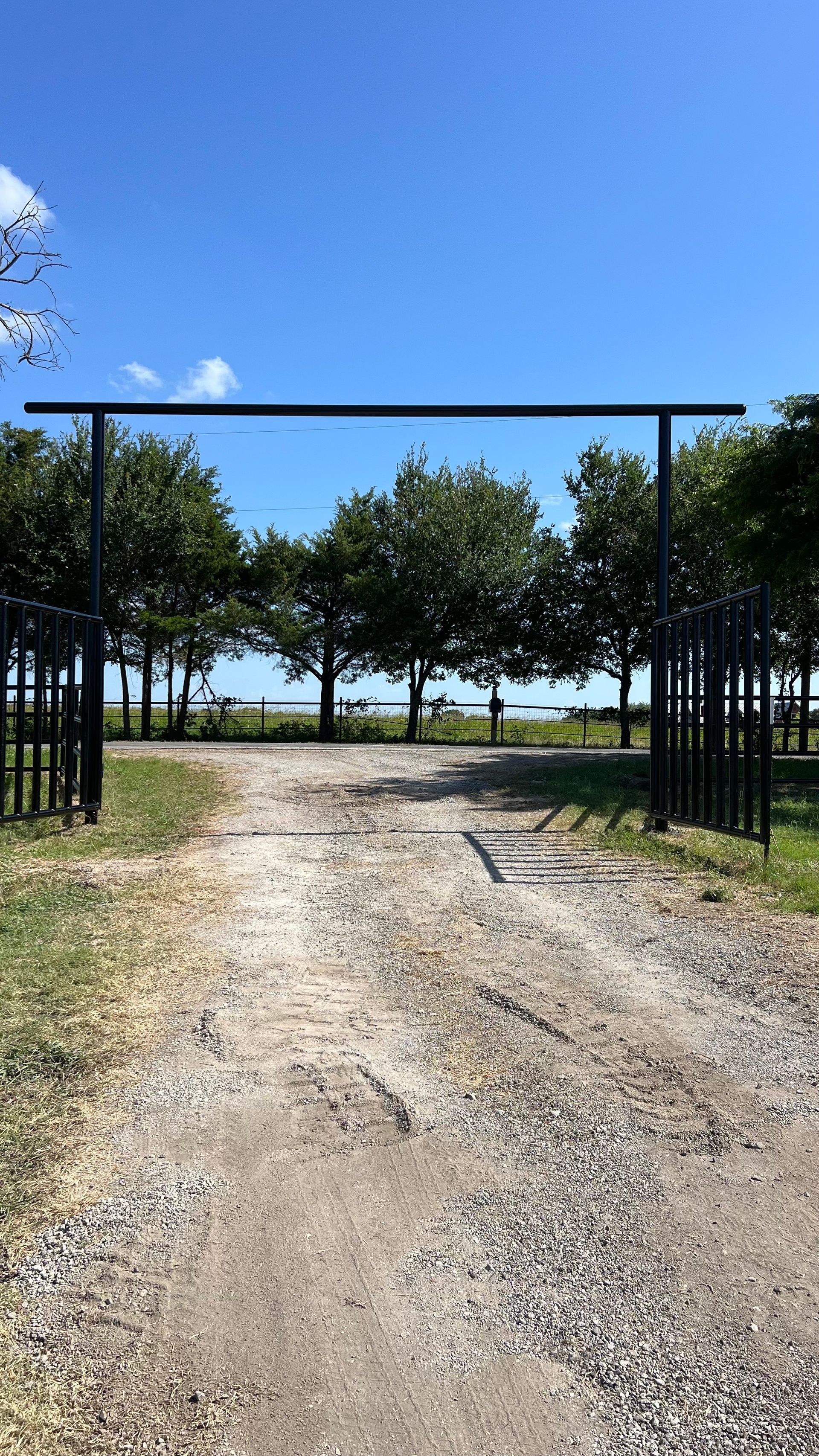 Gravel driveway leads to a black metal gate opening. Trees and water are visible beyond gate under blue sky.