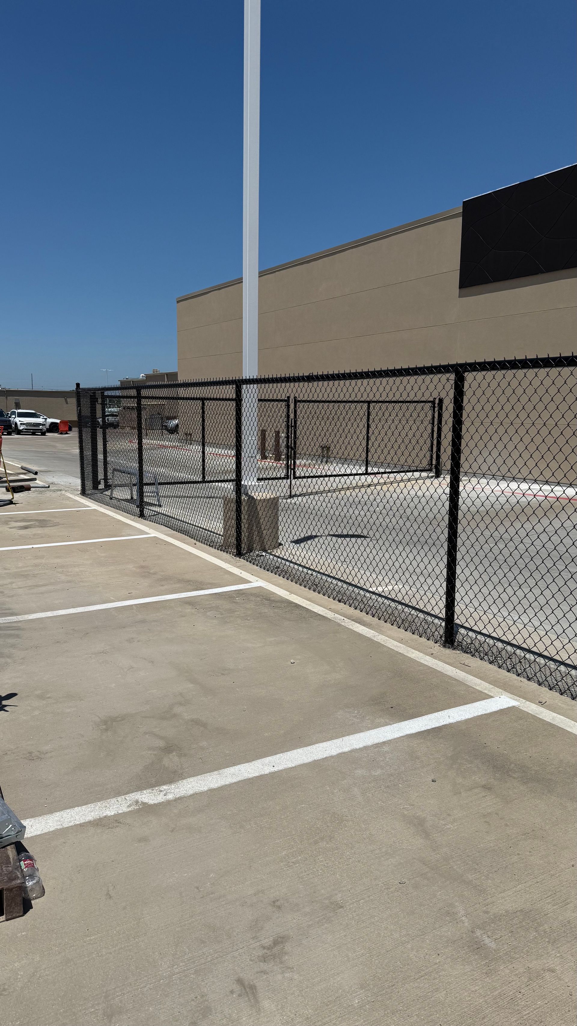 Black chain-link fence surrounds a gravel area near a building, with parking spaces marked by white lines.