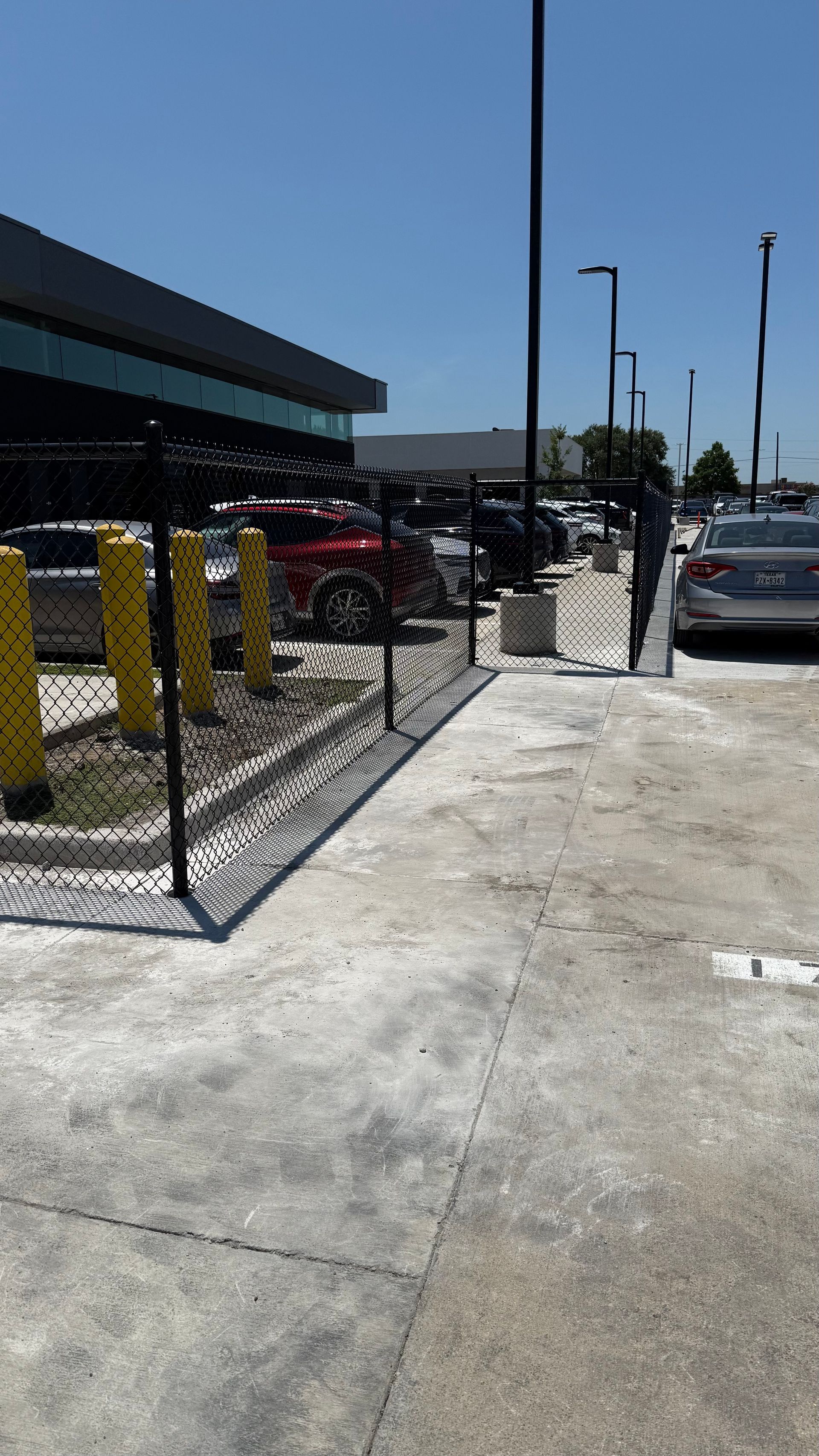 Black chain-link fence, concrete ground, and parked cars near a building under a bright blue sky.