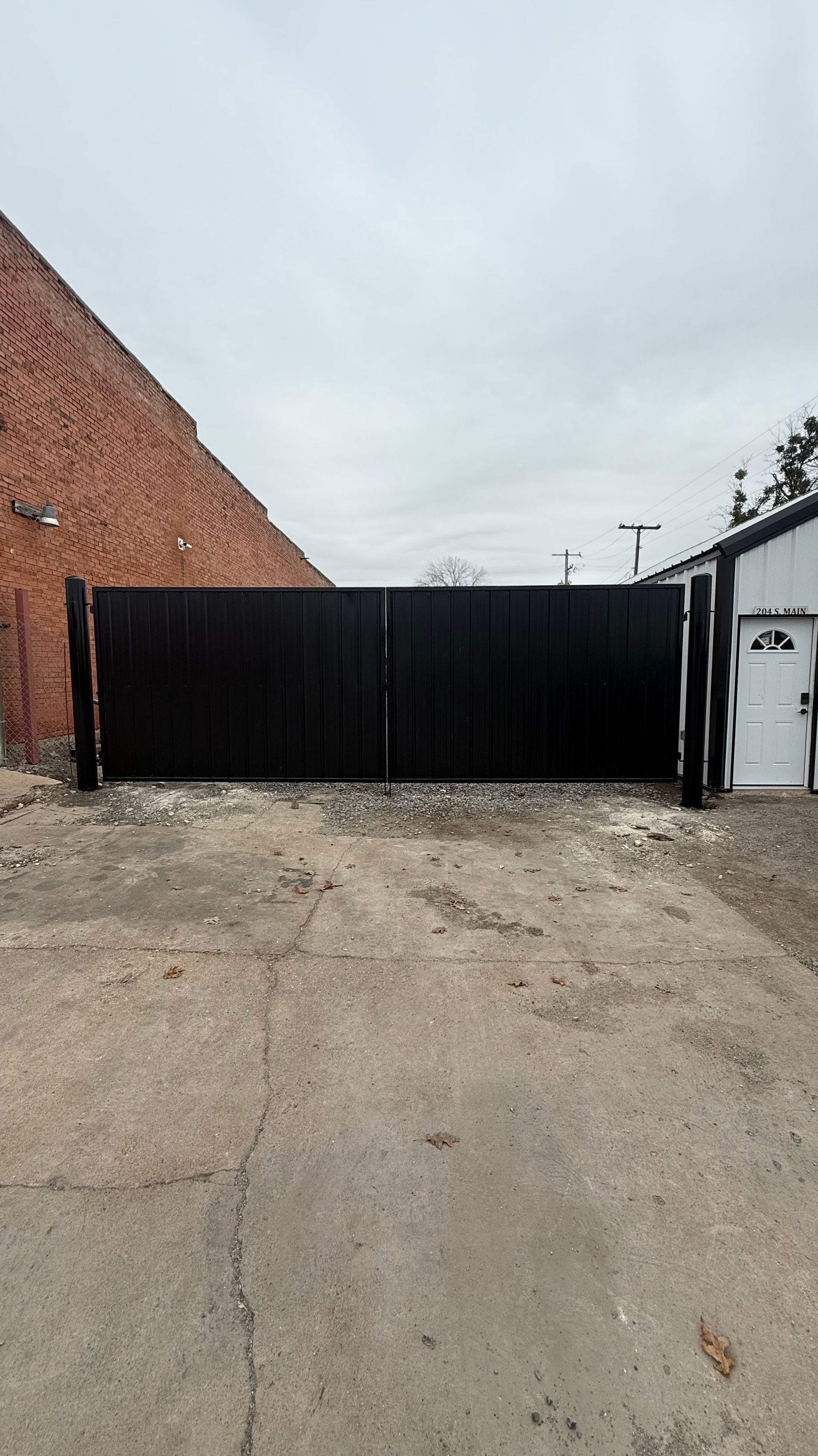 Black wooden double gate in front of a brick building and a white garage, on a paved lot.