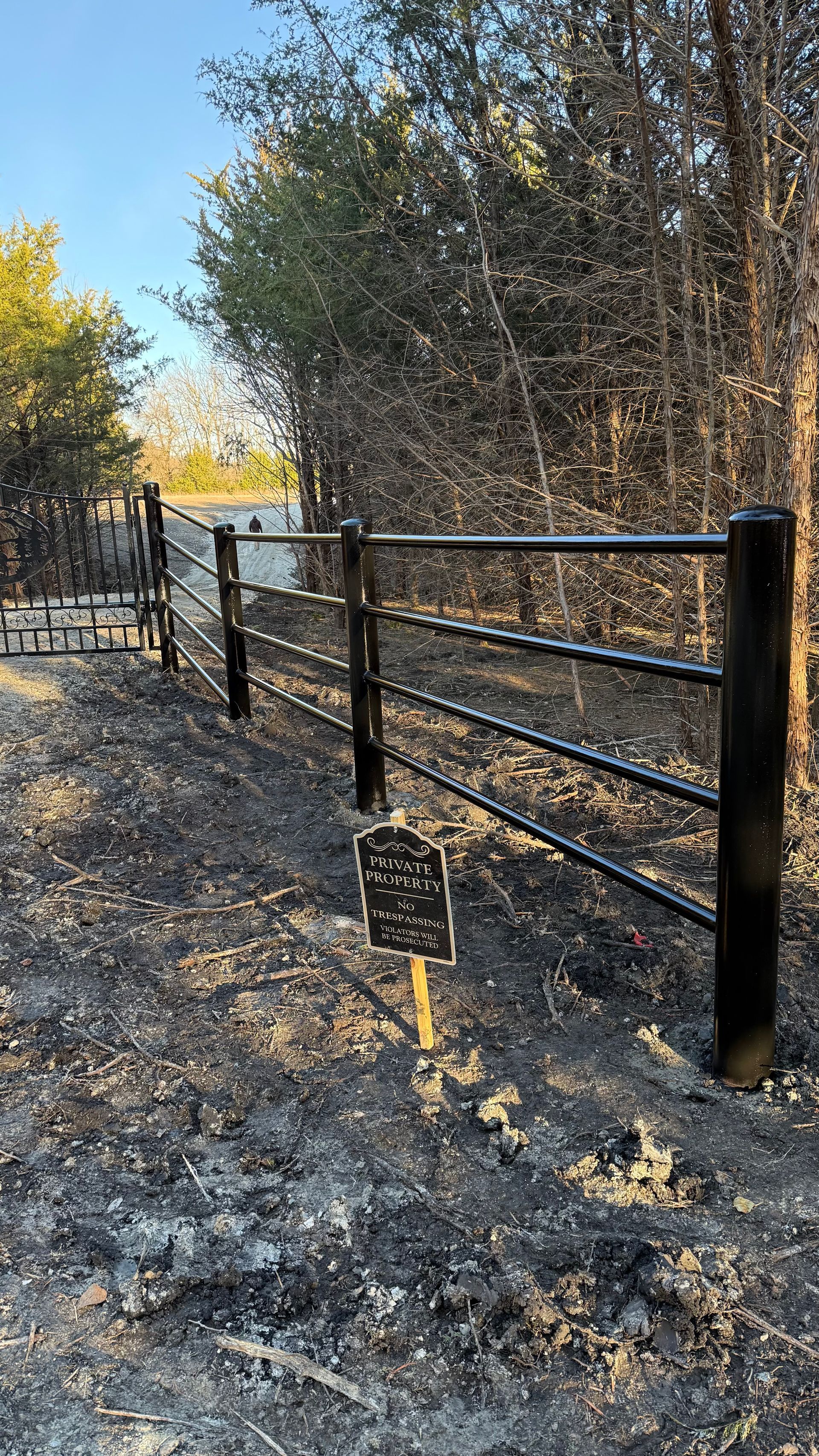 A black metal fence stands on scorched, ash-covered ground near a small, dark memorial sign in a wooded area.