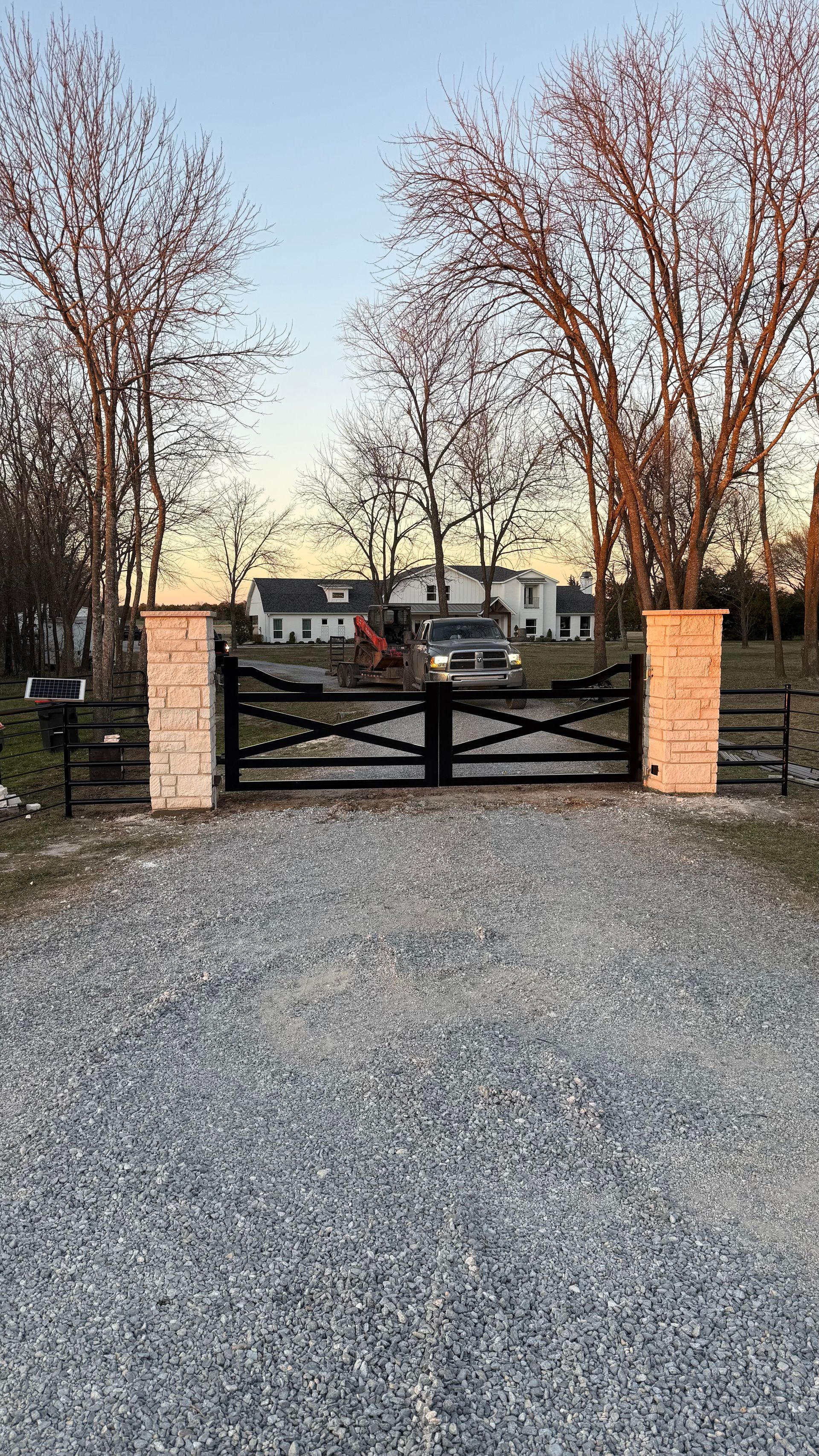 A gravel driveway leads to a closed black metal gate flanked by two tan stone pillars, with trees and a house beyond.