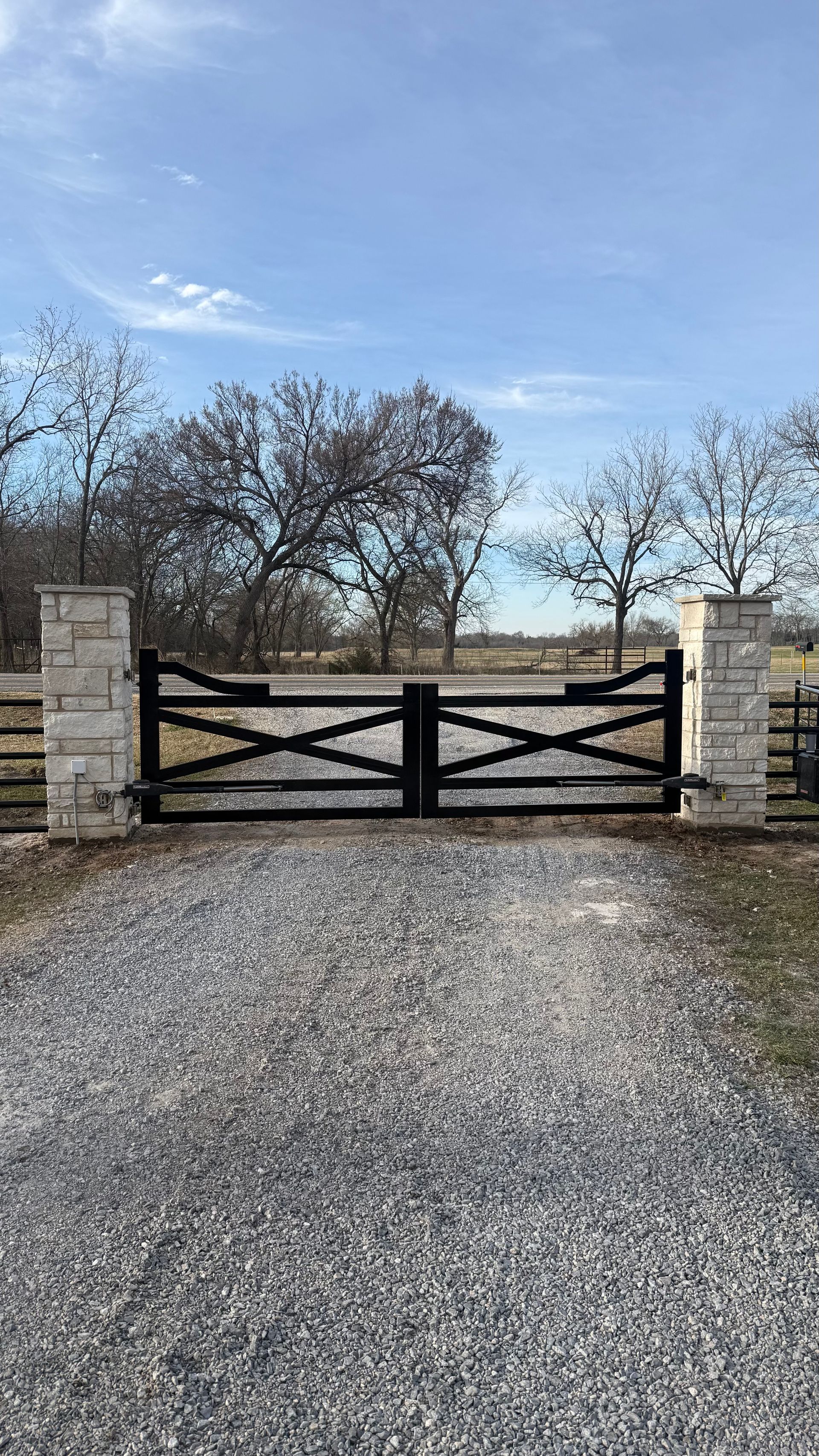 A black X-braced gate positioned between two light-colored stone pillars at the entrance of a gravel driveway.