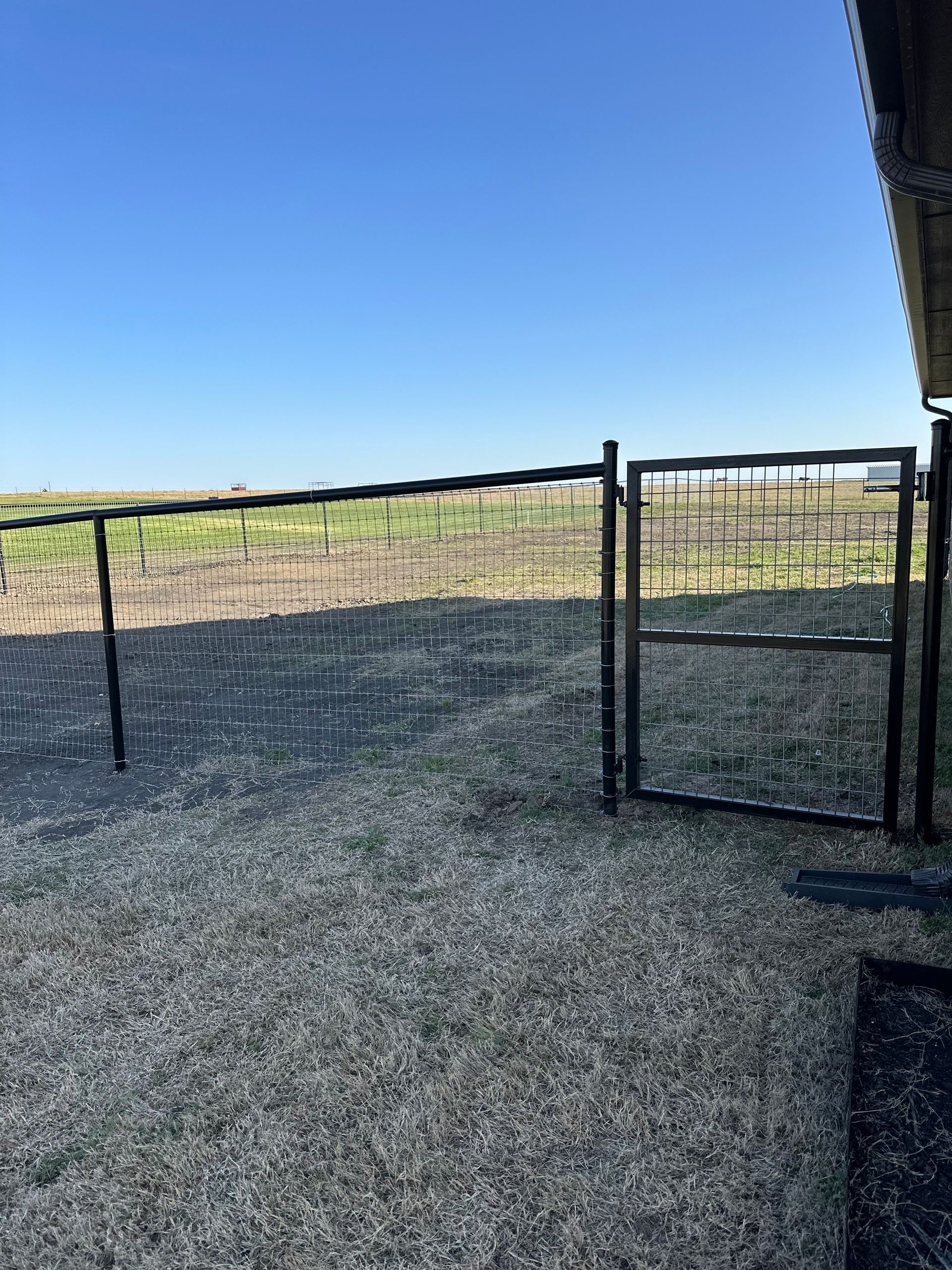 Black metal gate open in a gravel area, with a fence and field under a blue sky.