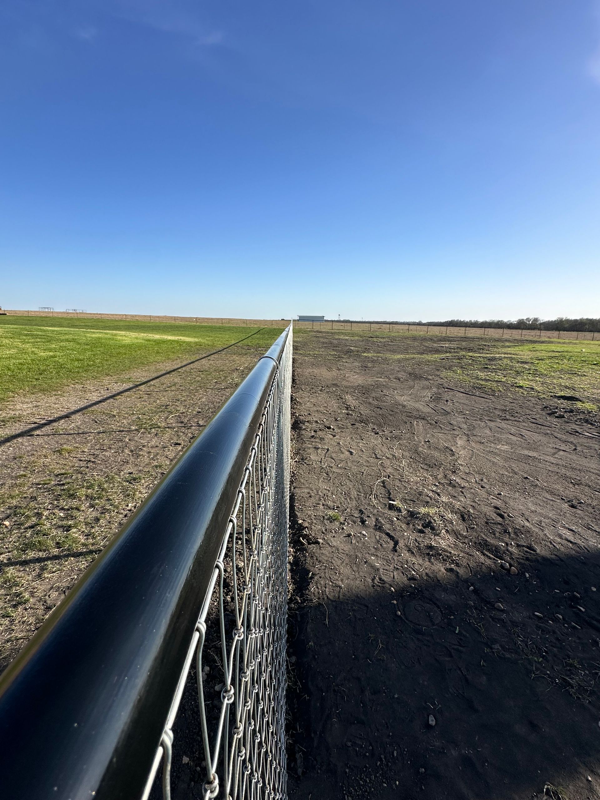 A black fence running along a gravel path, with fields on either side under a clear, blue sky.