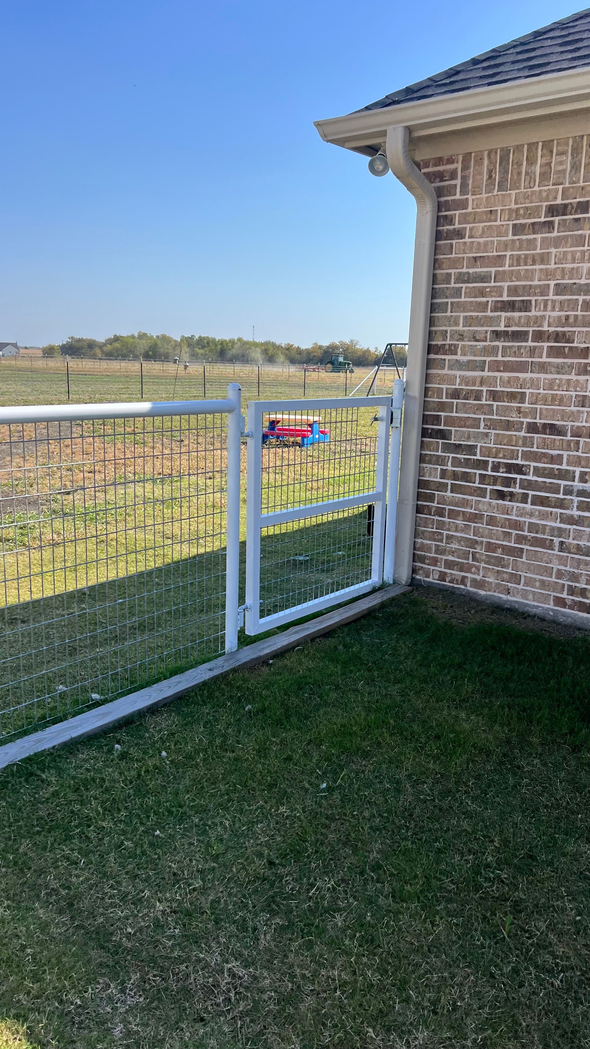 A white fence is surrounding a brick house with a field in the background.