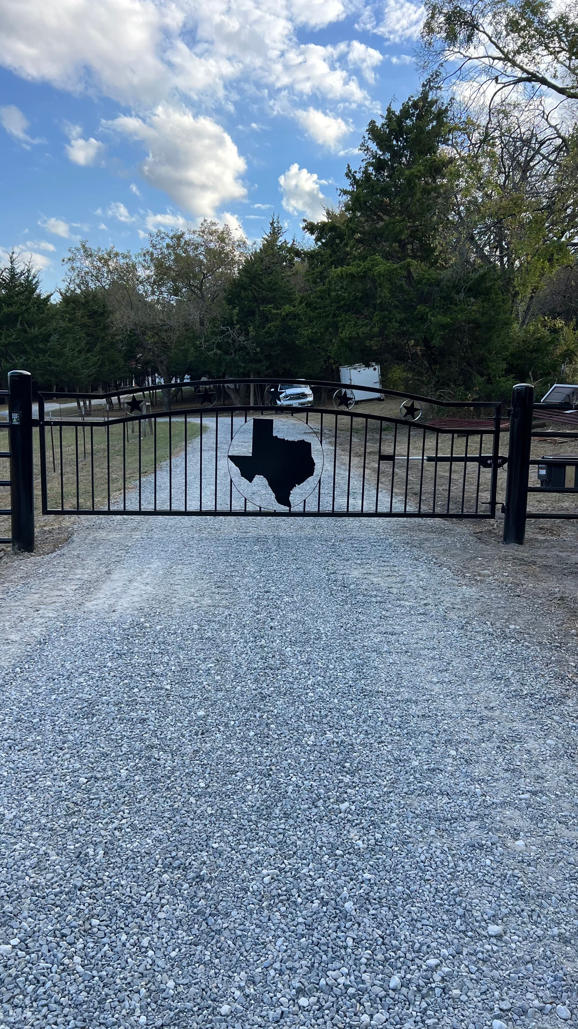 A black gate with a texas map on it is surrounded by gravel and trees.