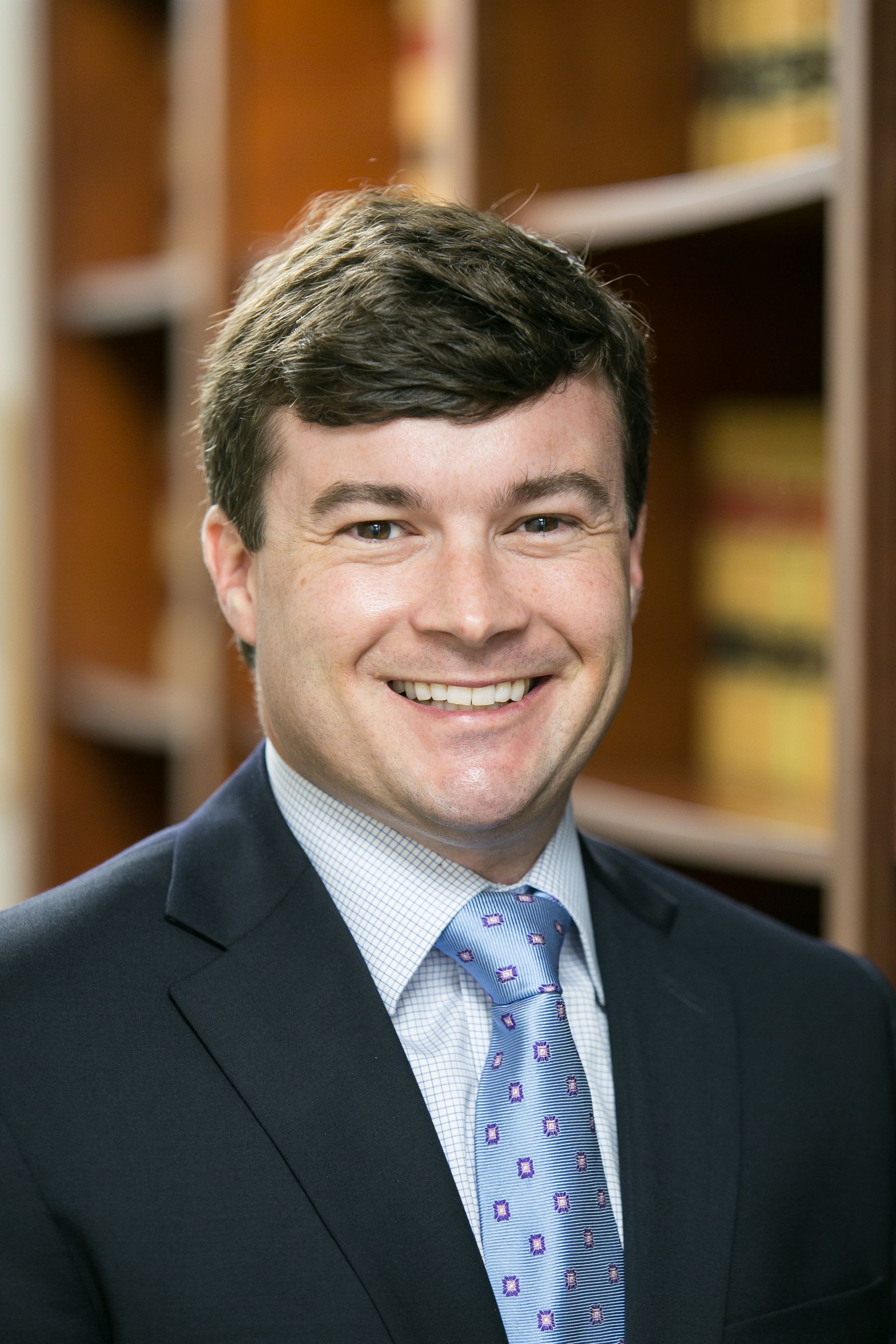Man in a suit smiles at the camera in an office setting with bookshelves.