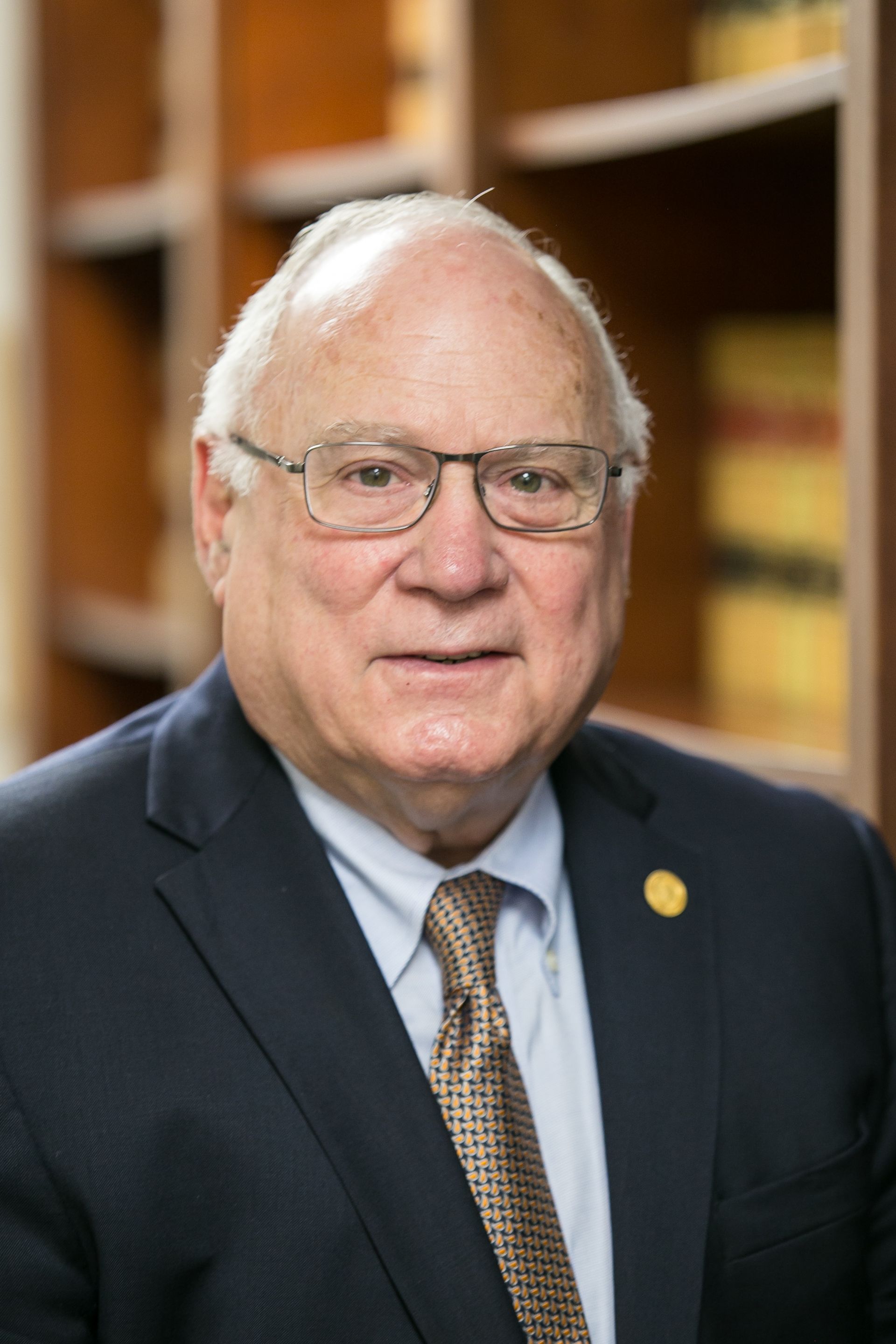 Older white man in a suit and glasses, smiles slightly, bookshelves in the background.