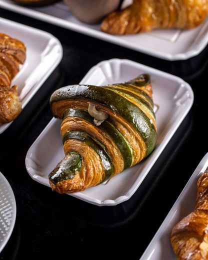 Croissant verde e oro a spirale, spolverato con zucchero a velo, su un vassoio bianco.