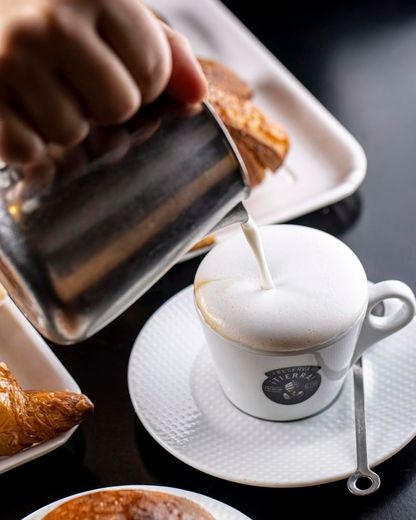 Latte versato in un cappuccino; croissant su un piatto lì vicino.