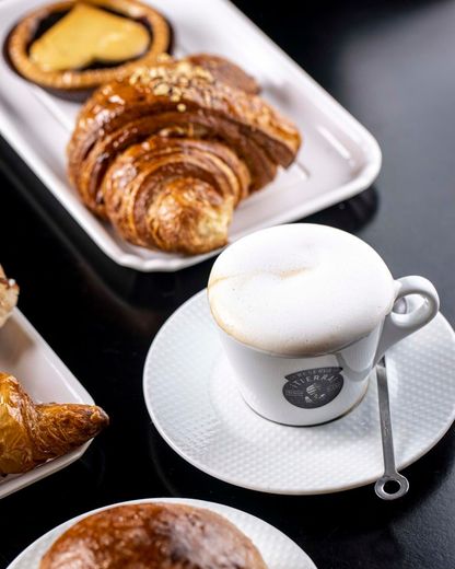 Cappuccino in tazza bianca con croissant e pasticcini su un tavolo nero.